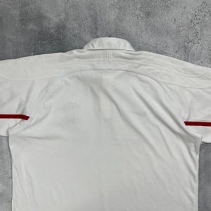 Pode incluir: Uma camisa branca com gola e detalhes vermelhos nas mangas. A camisa &eacute; feita de um tecido liso e tem um design cl&aacute;ssico. A camisa est&aacute; estendida sobre uma superf&iacute;cie cinza.