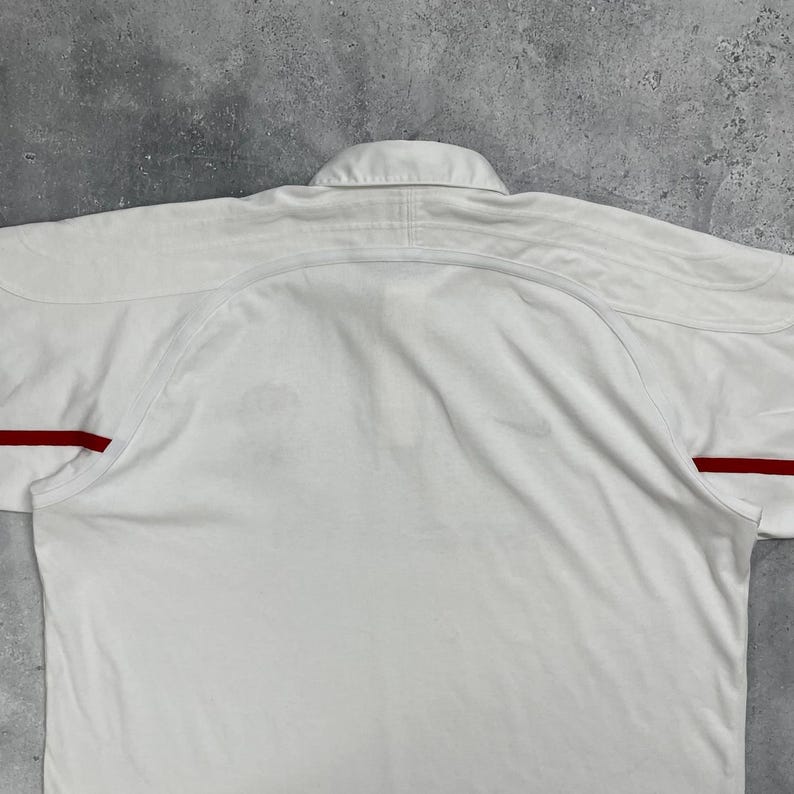 Pode incluir: Uma camisa branca com gola e detalhes vermelhos nas mangas. A camisa &eacute; feita de um tecido liso e tem um design cl&aacute;ssico. A camisa est&aacute; estendida sobre uma superf&iacute;cie cinza.