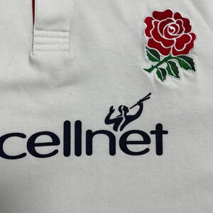 Pode incluir: Camisa polo branca com um emblema de rosa vermelha e a palavra "cellnet" em azul marinho. A camisa tem uma gola vermelha e uma carcela abotoada. Uma figura estilizada est&aacute; acima da palavra "cellnet".