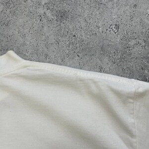 Peut inclure: Gros plan sur un t-shirt blanc, montrant l'encolure et la couture de l'&eacute;paule. Le tissu semble &ecirc;tre une mati&egrave;re douce, peut-&ecirc;tre du coton. Le fond est une surface grise mouchet&eacute;e.