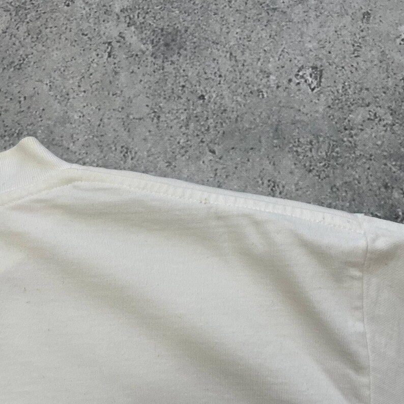 Peut inclure: Gros plan sur un t-shirt blanc, montrant l'encolure et la couture de l'&eacute;paule. Le tissu semble &ecirc;tre une mati&egrave;re douce, peut-&ecirc;tre du coton. Le fond est une surface grise mouchet&eacute;e.