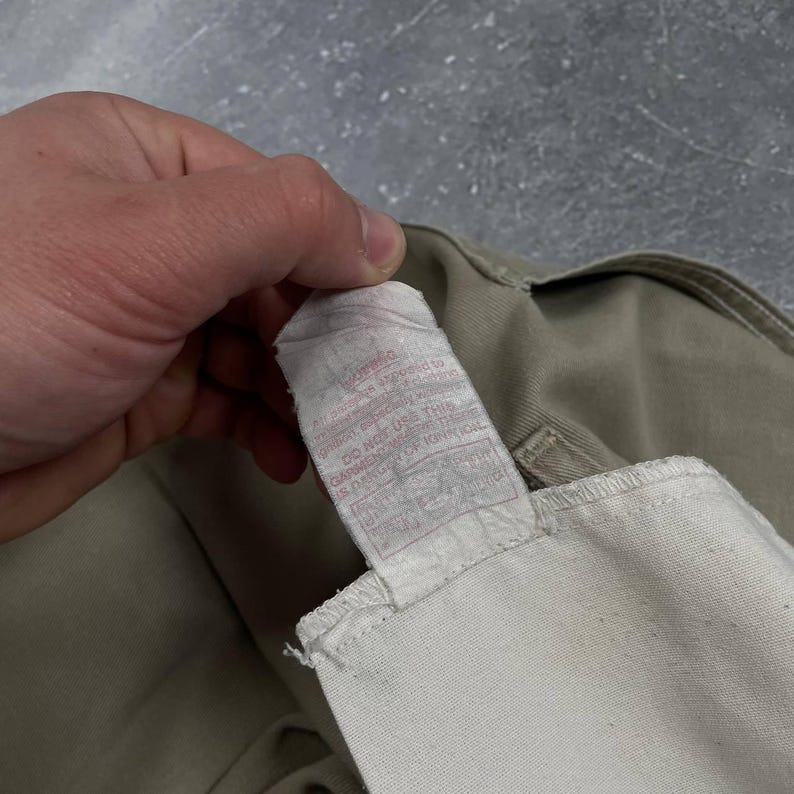 May include: Close-up of a beige garment tag with red text. The tag is attached to a beige fabric. The text on the tag is partially visible, with phrases like "DO NOT USE THIS GARMENT" and other warnings.