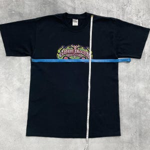 Op de afbeelding: Zwart T-shirt met een kleurrijke "Grave Digger" afbeelding over de borst. De afbeelding heeft een schedel- en vlammenontwerp met de tekst in een gestileerd lettertype. Een meetlint ligt over het shirt.