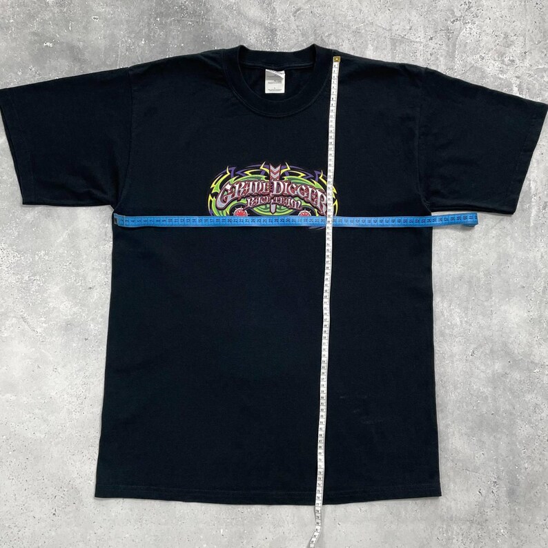Op de afbeelding: Zwart T-shirt met een kleurrijke "Grave Digger" afbeelding over de borst. De afbeelding heeft een schedel- en vlammenontwerp met de tekst in een gestileerd lettertype. Een meetlint ligt over het shirt.