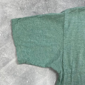 Pu&ograve; includere: Primo piano di una sezione di una maglietta verde, che mostra la manica e parte del corpo. Il tessuto ha un aspetto strutturato, con un effetto leggermente m&eacute;lange. Il colore &egrave; un verde tenue e morbido.