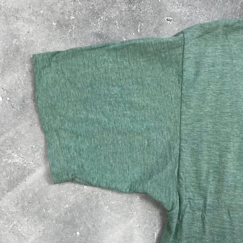 Pu&ograve; includere: Primo piano di una sezione di una maglietta verde, che mostra la manica e parte del corpo. Il tessuto ha un aspetto strutturato, con un effetto leggermente m&eacute;lange. Il colore &egrave; un verde tenue e morbido.