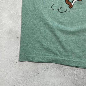 Pu&ograve; includere: T-shirt verde con grafica di un personaggio dei cartoni animati. Il personaggio &egrave; marrone e bianco. La parola "Cle" &egrave; scritta in corsivo con inchiostro nero.