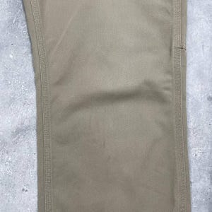 May include: Close-up of tan-colored pants. The pants have a straight leg design with visible stitching along the seams. The fabric has a subtle texture, and the overall look is casual and durable. The pants are a neutral color.