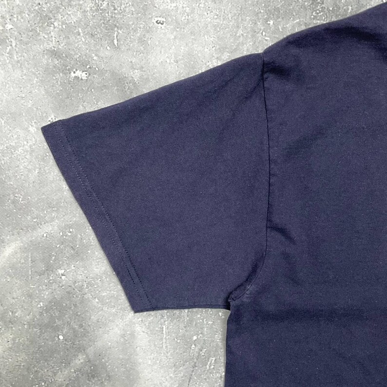 K&ouml;nnte beinhalten: Nahaufnahme eines marineblauen T-Shirt-&Auml;rmels. Der &Auml;rmel besteht aus einem weichen, undurchsichtigen Stoff und hat einen gen&auml;hten Saum. Das Shirt ist einfarbig und scheint ein Freizeitkleidungsst&uuml;ck zu sein.