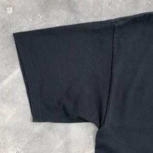 Op de afbeelding: Close-up van een zwarte t-shirtmouw. De mouw is gemaakt van een zachte, donkere stof en is langs de randen gestikt. Het t-shirt ligt op een grijze ondergrond.