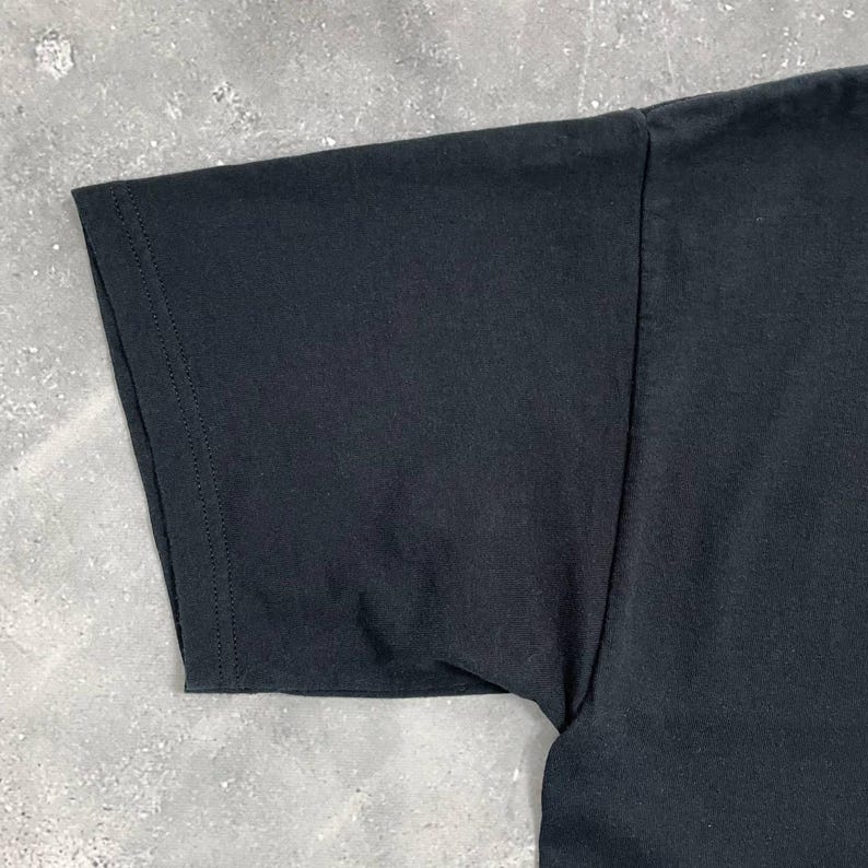 Op de afbeelding: Close-up van een zwarte t-shirtmouw. De mouw is gemaakt van een zachte, donkere stof en is langs de randen gestikt. Het t-shirt ligt op een grijze ondergrond.