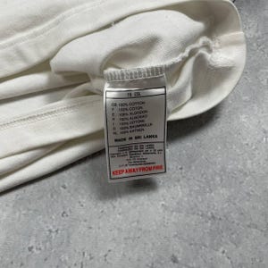 Pode incluir: Close-up de uma pe&ccedil;a de roupa de algod&atilde;o branca com uma etiqueta anexada. A etiqueta exibe texto em v&aacute;rios idiomas, incluindo "100% COTTON" e "MADE IN SRI LANKA". O tecido da pe&ccedil;a de roupa &eacute; vis&iacute;vel, com uma costura.