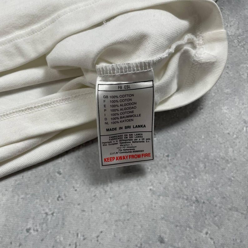 Pode incluir: Close-up de uma pe&ccedil;a de roupa de algod&atilde;o branca com uma etiqueta anexada. A etiqueta exibe texto em v&aacute;rios idiomas, incluindo "100% COTTON" e "MADE IN SRI LANKA". O tecido da pe&ccedil;a de roupa &eacute; vis&iacute;vel, com uma costura.
