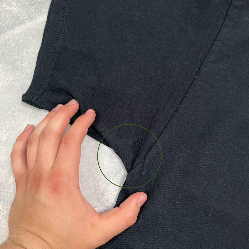 Op de afbeelding: Close-up van een zwart t-shirt, met de schouder- en mouwnaad. De stof is effen zwart van kleur en de stiksels zijn zichtbaar langs de naden. Het t-shirt lijkt gemaakt van een zacht materiaal.