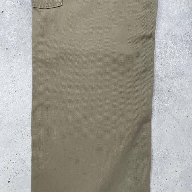 May include: Close-up of khaki-colored cargo pants. The pants have a pocket detail at the top and are made of a durable fabric. The color is a muted, earthy tone, suitable for outdoor or casual wear.