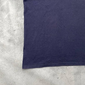 K&ouml;nnte beinhalten: Nahaufnahme eines marineblauen T-Shirts. Der Stoff scheint ein weiches Strickmaterial zu sein. Der untere Saum ist sichtbar, mit einer Doppelnaht. Das T-Shirt ist vor einem gesprenkelten grauen Hintergrund platziert.