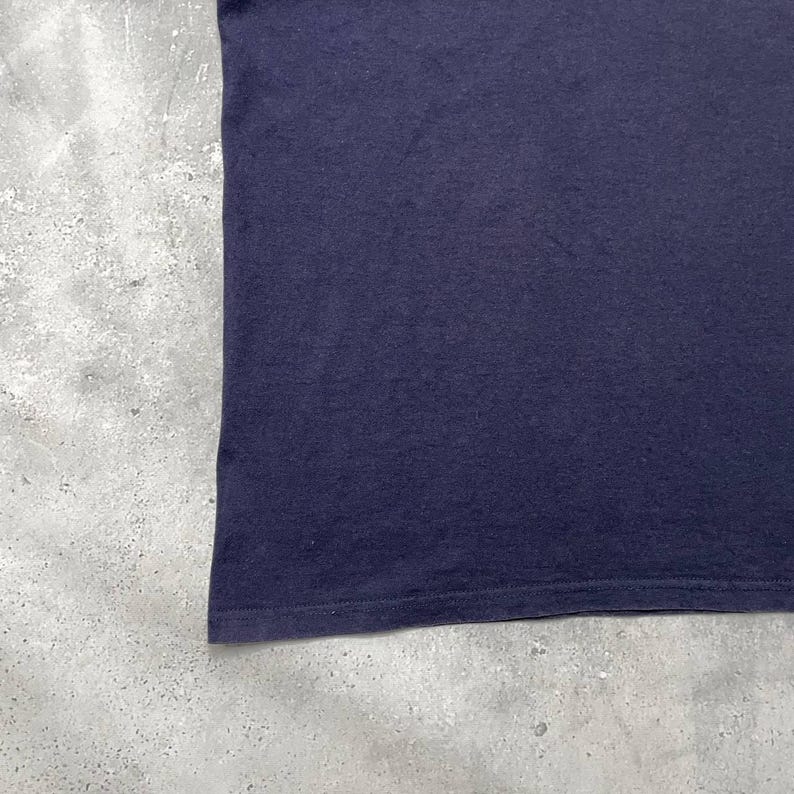 K&ouml;nnte beinhalten: Nahaufnahme eines marineblauen T-Shirts. Der Stoff scheint ein weiches Strickmaterial zu sein. Der untere Saum ist sichtbar, mit einer Doppelnaht. Das T-Shirt ist vor einem gesprenkelten grauen Hintergrund platziert.