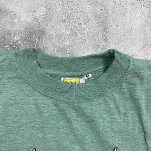 Pu&ograve; includere: Primo piano di una maglietta verde con scollo tondo. La maglietta ha un'etichetta con la scritta "best" e un'etichetta con la taglia "M". La maglietta &egrave; realizzata in un materiale morbido.