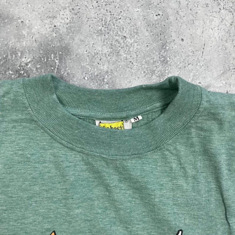 Pu&ograve; includere: Primo piano di una maglietta verde con scollo tondo. La maglietta ha un'etichetta con la scritta "best" e un'etichetta con la taglia "M". La maglietta &egrave; realizzata in un materiale morbido.