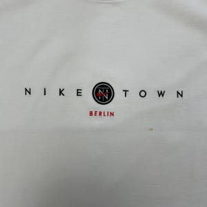 Peut inclure: T-shirt blanc &agrave; col rond avec le texte "NIKE TOWN" en noir. Le mot "BERLIN" est en rouge sous un logo circulaire avec "N" et "T" &agrave; l'int&eacute;rieur. Le t-shirt est de style d&eacute;contract&eacute;.