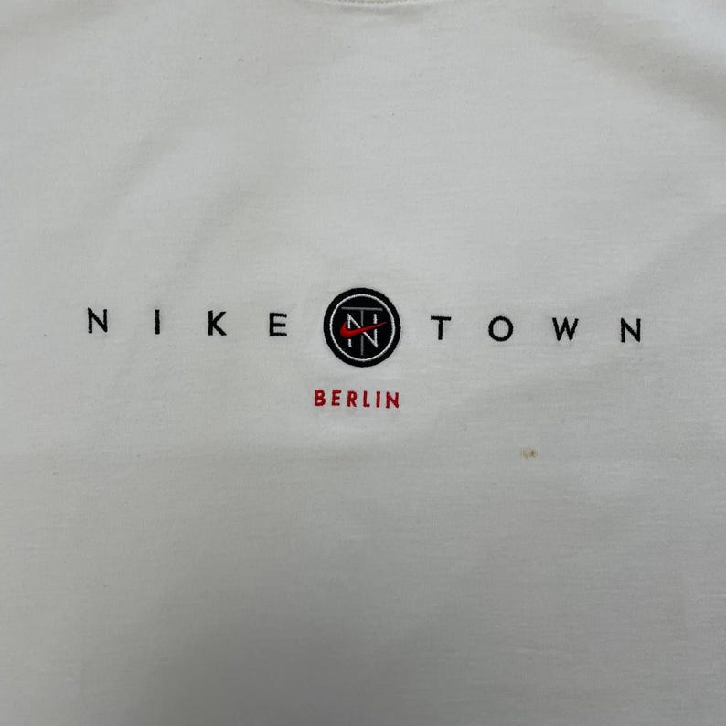 Peut inclure: T-shirt blanc &agrave; col rond avec le texte "NIKE TOWN" en noir. Le mot "BERLIN" est en rouge sous un logo circulaire avec "N" et "T" &agrave; l'int&eacute;rieur. Le t-shirt est de style d&eacute;contract&eacute;.
