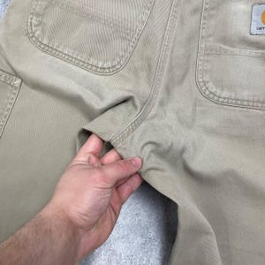 May include: Close-up of tan work pants with multiple pockets and reinforced stitching. The Carhartt logo is visible on the back pocket. The pants are made of a durable, heavy-duty fabric.