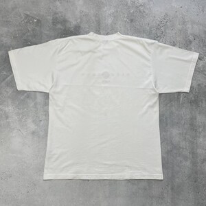 Peut inclure: Un t-shirt blanc &agrave; col rond et manches courtes. Le t-shirt porte un motif d&eacute;lav&eacute; sur le haut du dos. Le t-shirt est fait d'un tissu doux et l&eacute;ger.