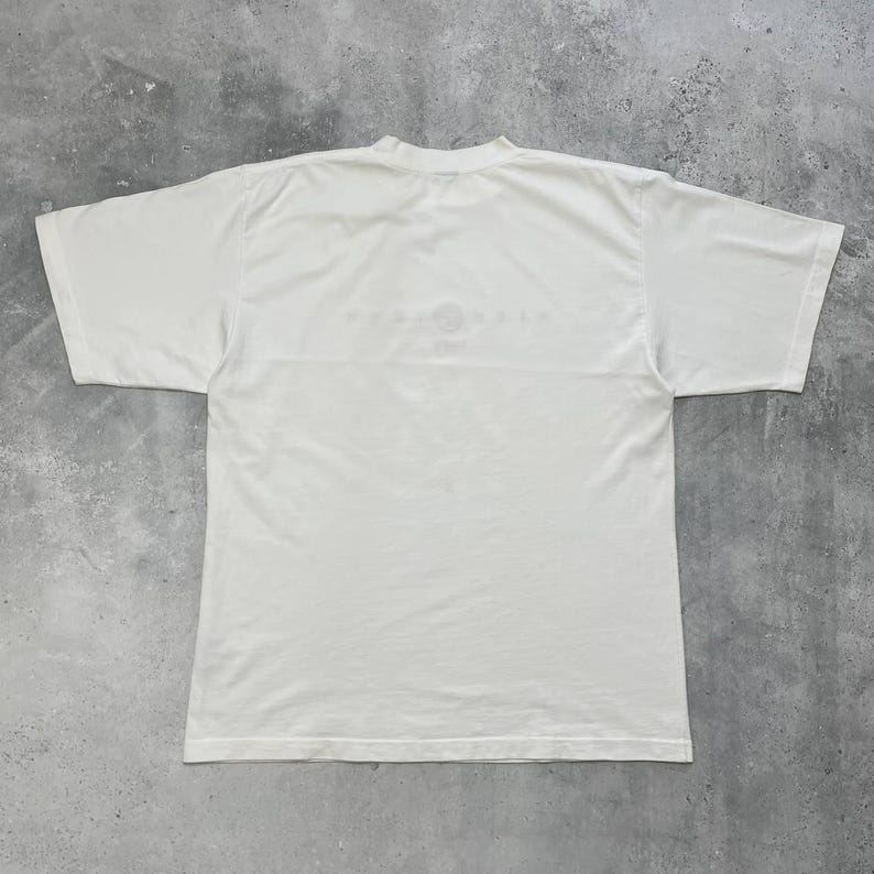 Peut inclure: Un t-shirt blanc &agrave; col rond et manches courtes. Le t-shirt porte un motif d&eacute;lav&eacute; sur le haut du dos. Le t-shirt est fait d'un tissu doux et l&eacute;ger.