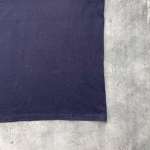 K&ouml;nnte beinhalten: Nahaufnahme eines dunkelmarineblauen T-Shirts. Der Stoff wirkt weich und leicht strukturiert. Der untere Saum des Shirts ist sichtbar, mit einer feinen Naht. Der Hintergrund ist eine hellgraue, strukturierte Oberfl&auml;che.