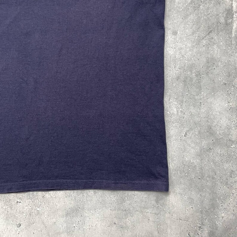K&ouml;nnte beinhalten: Nahaufnahme eines dunkelmarineblauen T-Shirts. Der Stoff wirkt weich und leicht strukturiert. Der untere Saum des Shirts ist sichtbar, mit einer feinen Naht. Der Hintergrund ist eine hellgraue, strukturierte Oberfl&auml;che.
