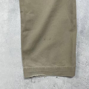 May include: Close-up of the bottom of a pair of khaki-colored pants. The fabric is a solid color with a slightly textured appearance. The hem of the pants is visible, with some fraying at the edge. The pants are on a gray surface.