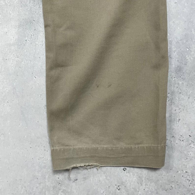 May include: Close-up of the bottom of a pair of khaki-colored pants. The fabric is a solid color with a slightly textured appearance. The hem of the pants is visible, with some fraying at the edge. The pants are on a gray surface.