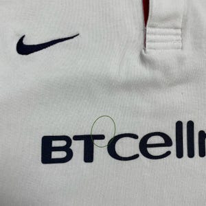 Pode incluir: Camiseta esportiva branca com o logotipo da Nike em azul marinho e o texto "BTcell" em azul marinho. A camisa tem um detalhe vermelho na gola.