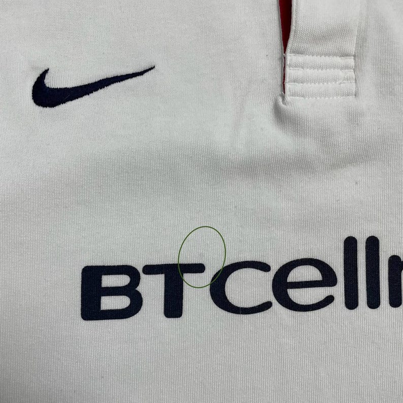 Pode incluir: Camiseta esportiva branca com o logotipo da Nike em azul marinho e o texto "BTcell" em azul marinho. A camisa tem um detalhe vermelho na gola.