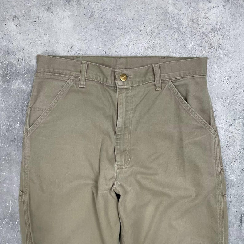 May include: Close-up of khaki-colored work pants. The pants have a button closure, belt loops, and multiple pockets. The fabric appears to be a durable cotton blend. The pants are designed for work or casual wear.
