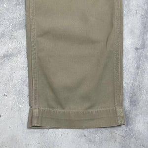 May include: Close-up of the bottom of khaki-colored pants. The fabric is a solid, neutral tone with visible stitching along the seams and hem. The pants appear to be made of a durable material, suitable for work or casual wear.