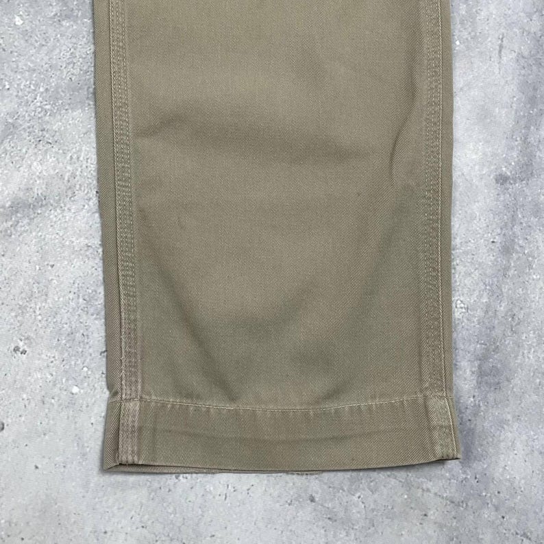 May include: Close-up of the bottom of khaki-colored pants. The fabric is a solid, neutral tone with visible stitching along the seams and hem. The pants appear to be made of a durable material, suitable for work or casual wear.