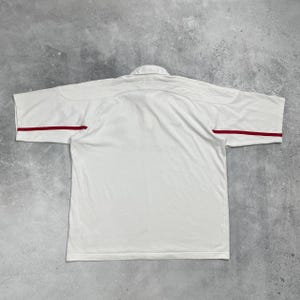 Pode incluir: Camisa polo branca com mangas curtas e gola. A camisa tem listras vermelhas nas mangas. A camisa &eacute; feita de um material leve.