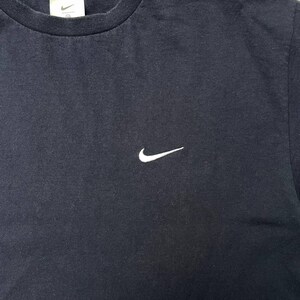 K&ouml;nnte beinhalten: Ein marineblaues T-Shirt mit einem kleinen wei&szlig;en Nike Swoosh-Logo auf der linken Brust. Das Shirt hat einen Rundhalsausschnitt und kurze &Auml;rmel. Der Stoff scheint eine weiche Baumwollmischung zu sein.