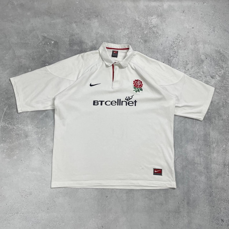 Pode incluir: Camisola de rugby branca de manga curta com gola. Apresenta um emblema de rosa vermelha e o texto "BTcellnet" em preto. Um logotipo da Nike preto &eacute; vis&iacute;vel no peito. A gola tem um detalhe vermelho.