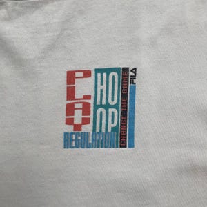 May include: White t-shirt with a colorful graphic design. The design features the words "PLAY," "HOOP," and "REGULATION" in red, teal, and blue, with the text "CHANGE THE GAME" and the brand name "FILA" in black and blue.