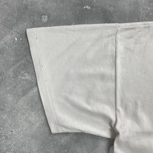 May include: Close-up of a section of a white t-shirt. The fabric is slightly wrinkled and shows a few small holes near the sleeve seam. The shirt appears to be made of a soft, lightweight material.