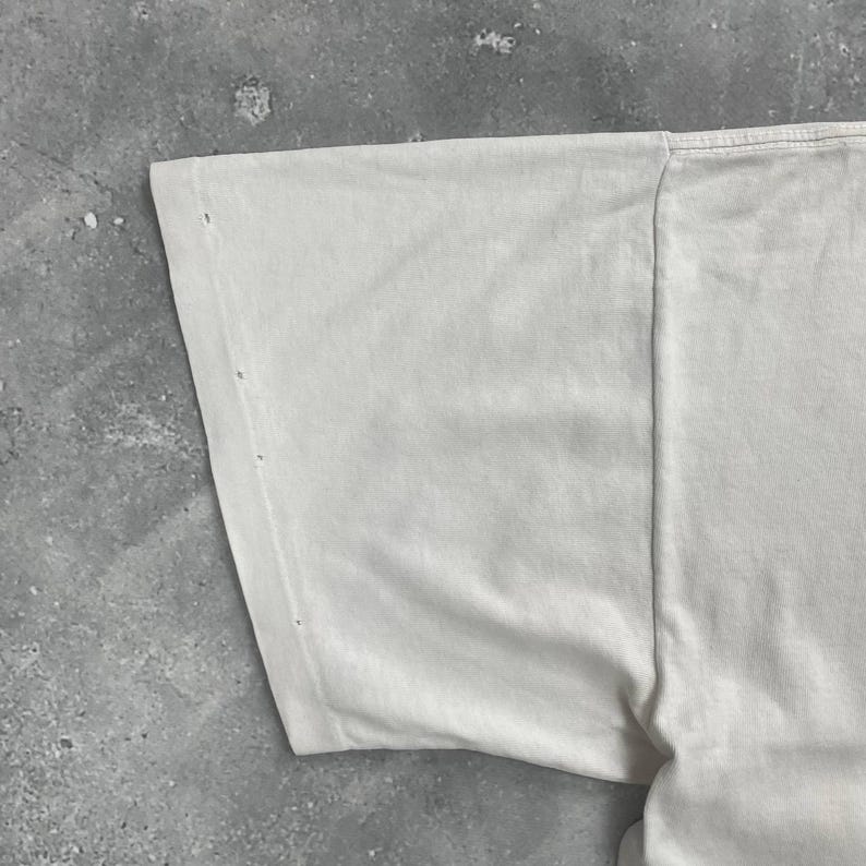 May include: Close-up of a section of a white t-shirt. The fabric is slightly wrinkled and shows a few small holes near the sleeve seam. The shirt appears to be made of a soft, lightweight material.