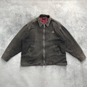 May include: A dark brown jacket with a full-length zipper, two front pockets, and a red and black plaid lining visible at the collar. A small red logo is on the upper left chest. The jacket is made of a durable material.