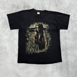Könnte beinhalten: Schwarzes T-Shirt mit einem verblichenen Grafikdruck einer Person und dem Bandnamen "Korn" in einer stilisierten Schriftart. Die Grafik ist in Braun- und Beigetönen gehalten und hat eine Vintage-Ästhetik. Das Shirt hat einen Rundhalsausschnitt und kurze Ärmel.