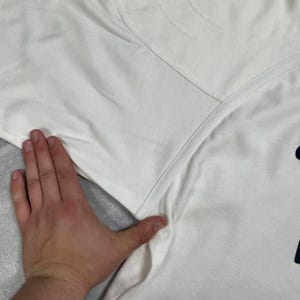 Pode incluir: Camiseta branca com gola redonda e um gr&aacute;fico azul marinho na parte inferior direita. A camiseta est&aacute; sendo segurada por uma m&atilde;o. O tecido parece ser algod&atilde;o.