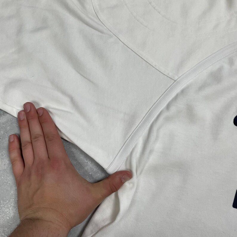 Pode incluir: Camiseta branca com gola redonda e um gr&aacute;fico azul marinho na parte inferior direita. A camiseta est&aacute; sendo segurada por uma m&atilde;o. O tecido parece ser algod&atilde;o.