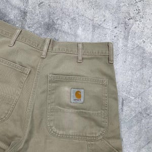 May include: Close-up of tan work pants with a Carhartt logo patch on the back pocket. The pants have a classic five-pocket design with reinforced stitching and a durable, workwear aesthetic. The fabric appears to be a sturdy cotton blend.