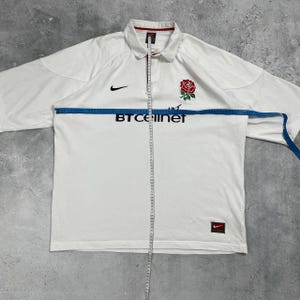Pode incluir: Camisa de rugby branca com um emblema de rosa vermelha e o texto "BT Cellnet". Apresenta um logotipo da Nike e uma gola. A camisa est&aacute; sendo medida com uma fita m&eacute;trica azul.
