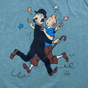 Pu&ograve; includere: T-shirt verde acqua con un'illustrazione a cartoni animati di due personaggi che si abbracciano. Uno indossa un abito e un cappello neri, l'altro una camicia blu e pantaloni marroni. Entrambi corrono con un bastone. Stelle e linee ondulate sparse.